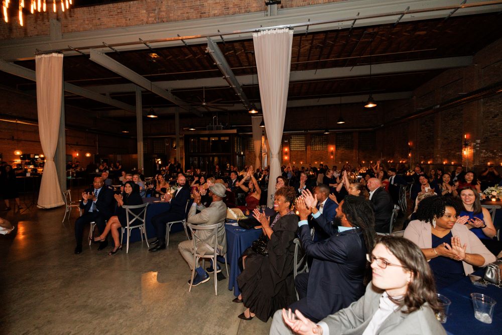 Attendees seated at long wooden square tables in a loft style event space, facing a stage adorned with elegant décor. They watch attentively as presenters announce award recipients during the gala
